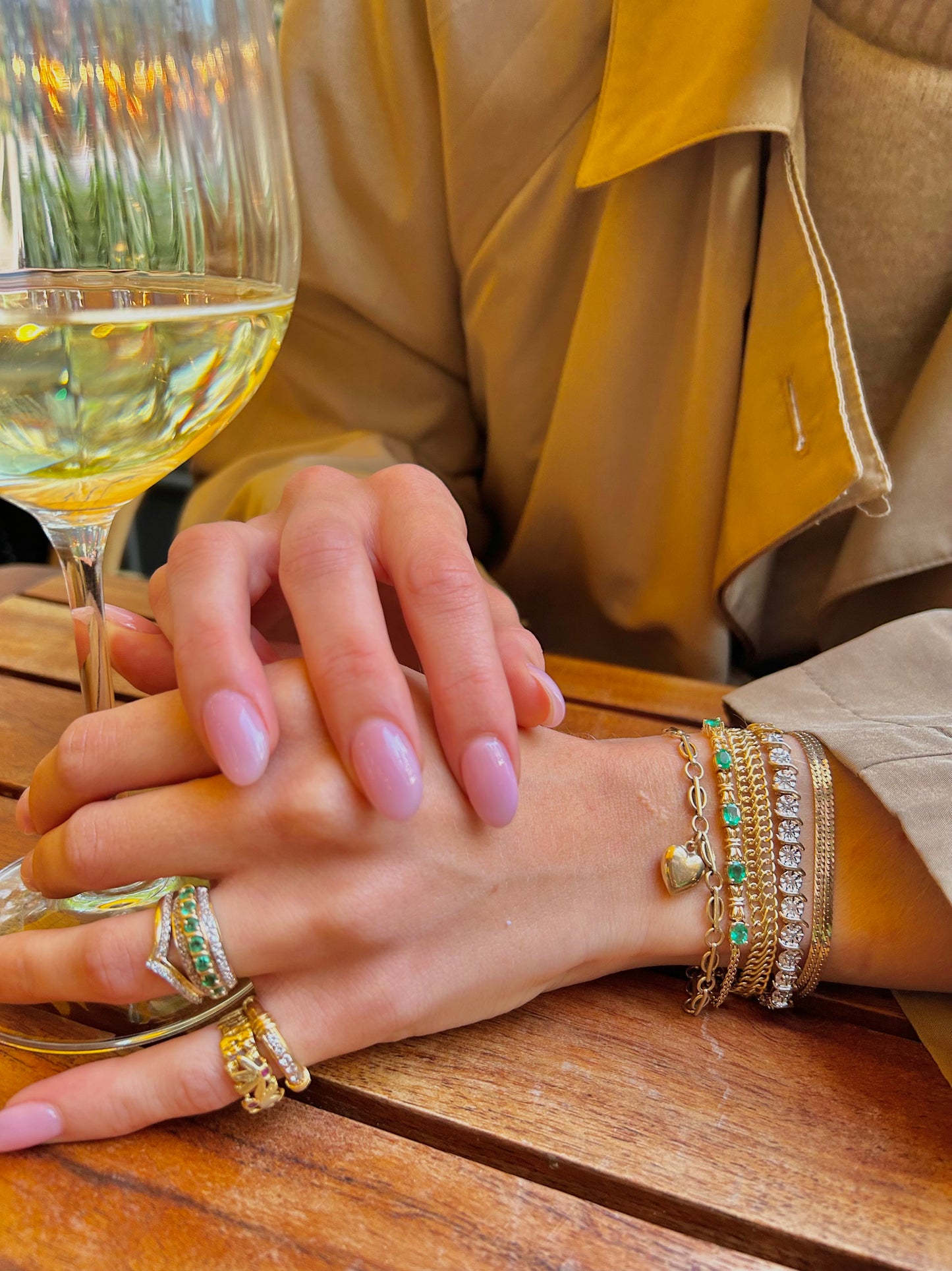 Close-up of a hand with pink nail polish and gold jewelry near a glass of white wine.