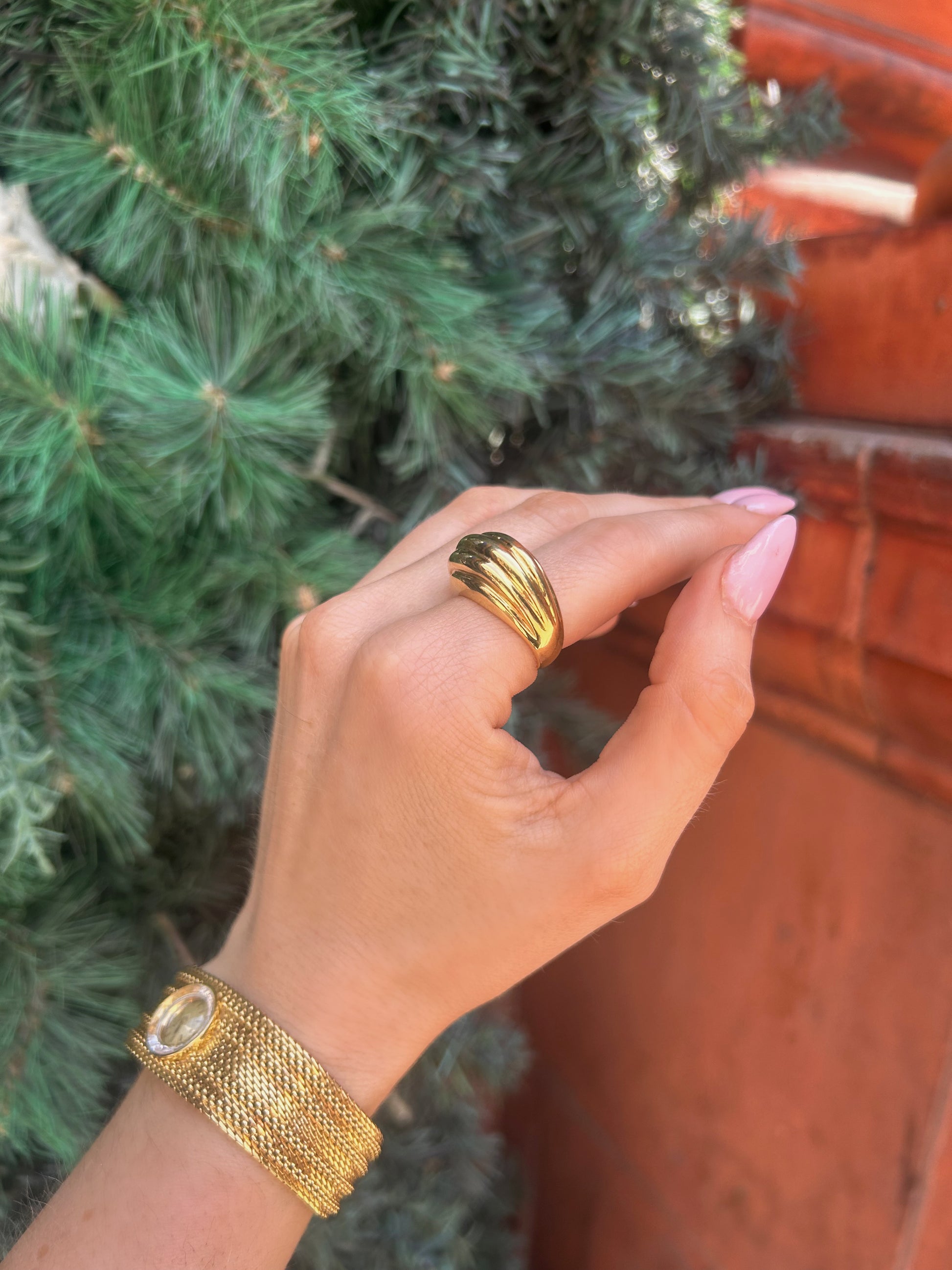 A close-up of a hand wearing a wide, multi-band gold ring and a matching gold mesh bangle bracelet, set against a background of green pine needles and a terracotta wall.