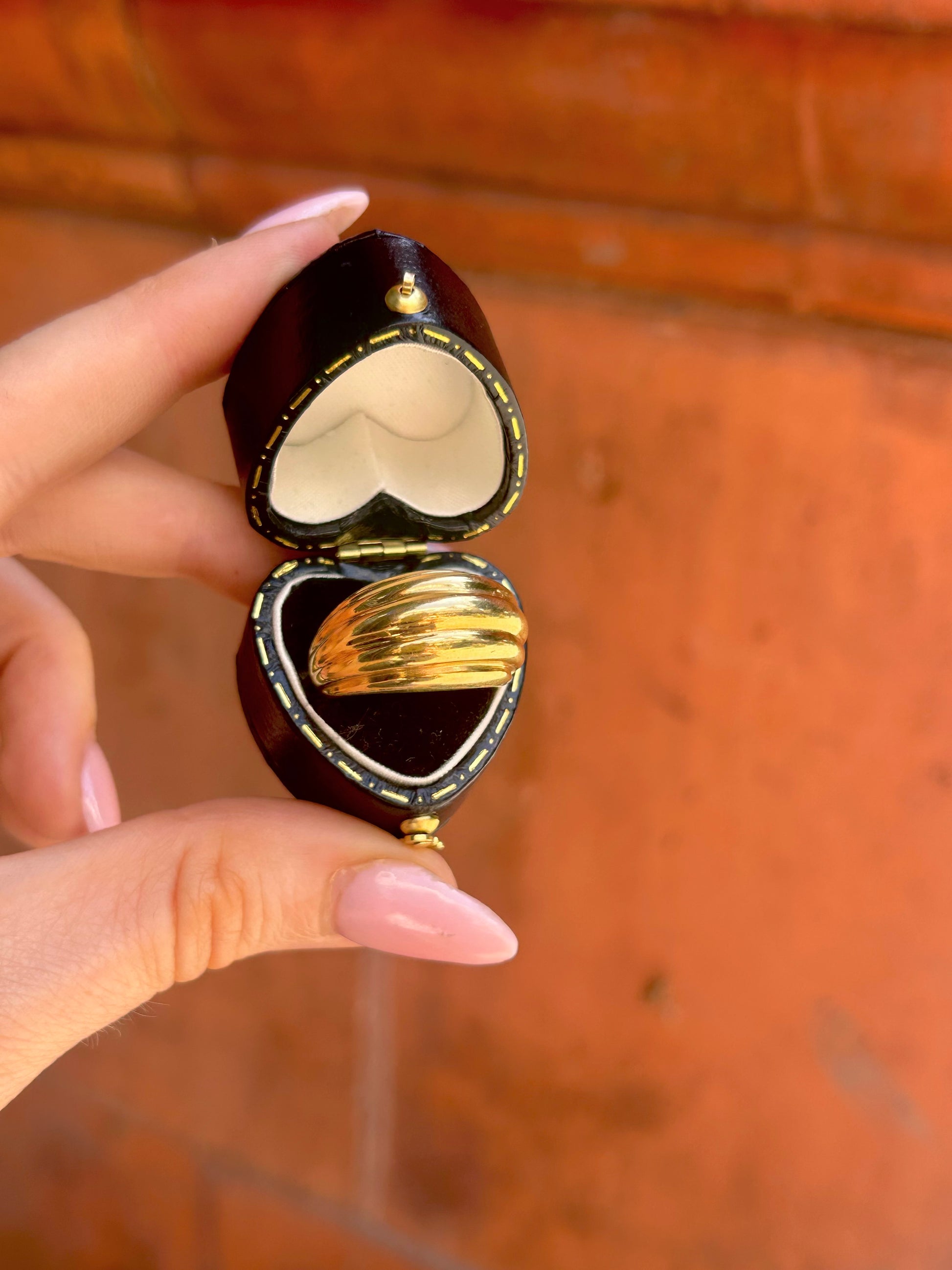 A hand with pink manicured nails holds an open heart-shaped dark brown wooden jewelry box containing two gold rings, set against a warm terracotta brick background.